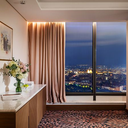 Grand Hotel Millennium โซเฟีย ภายนอก รูปภาพ The photo depicts a modern hotel room or a lounge area with a stunning view of a cityscape at night. On the left, there's a wooden console table with a vase of flowers and a bottle of water. A piece of art is framed on the wall above the table. The r