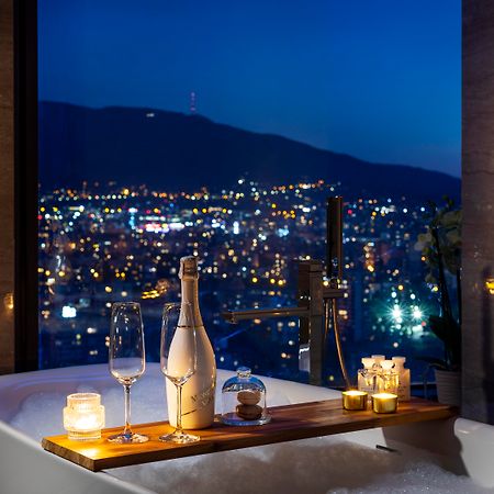 Grand Hotel Millennium โซเฟีย ภายนอก รูปภาพ The photo features a luxurious bathtub placed by a large window, showcasing a scenic city view at night. There is a wooden tray resting on the edge of the tub, displaying a bottle of champagne, two glass flutes, and several lit candles, creating an a