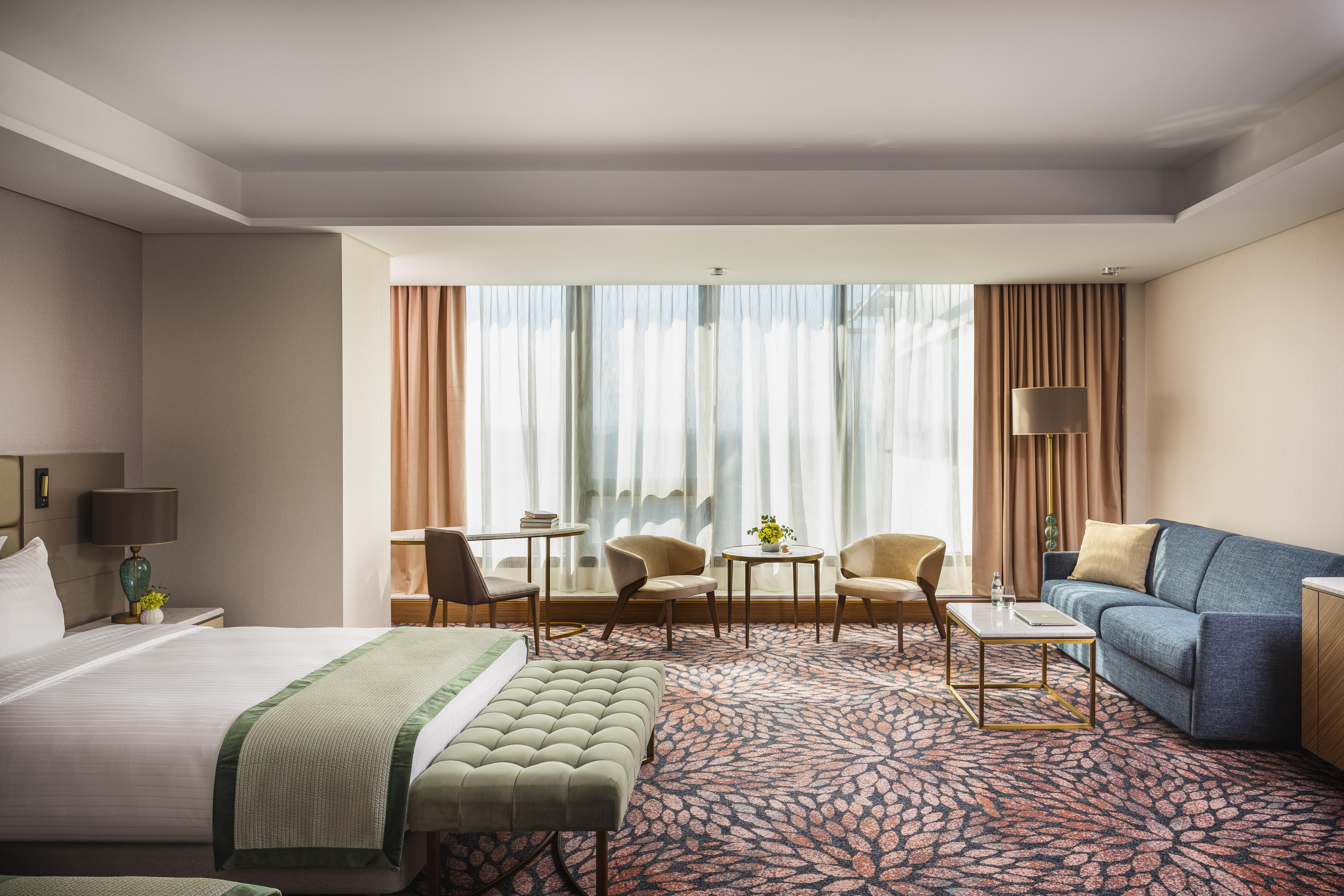 Grand Hotel Millennium โซเฟีย ภายนอก รูปภาพ The photo shows a modern hotel room featuring a spacious layout. On one side, there is a large bed with a green throw blanket and a tufted bench at its foot. The room is well-lit by large windows dressed with light curtains, which allow natural light
