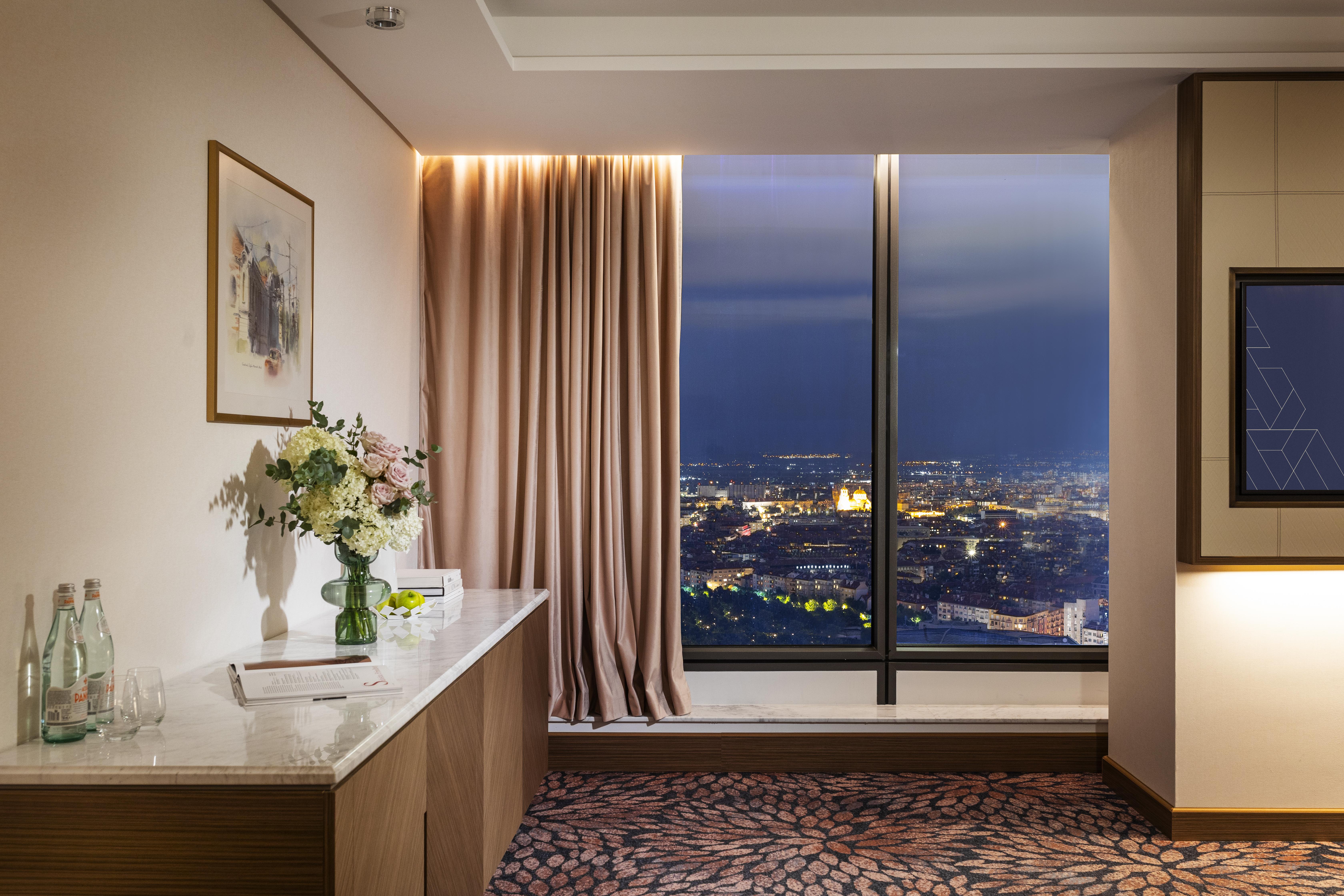 Grand Hotel Millennium โซเฟีย ภายนอก รูปภาพ The photo depicts a modern hotel room or a lounge area with a stunning view of a cityscape at night. On the left, there's a wooden console table with a vase of flowers and a bottle of water. A piece of art is framed on the wall above the table. The r