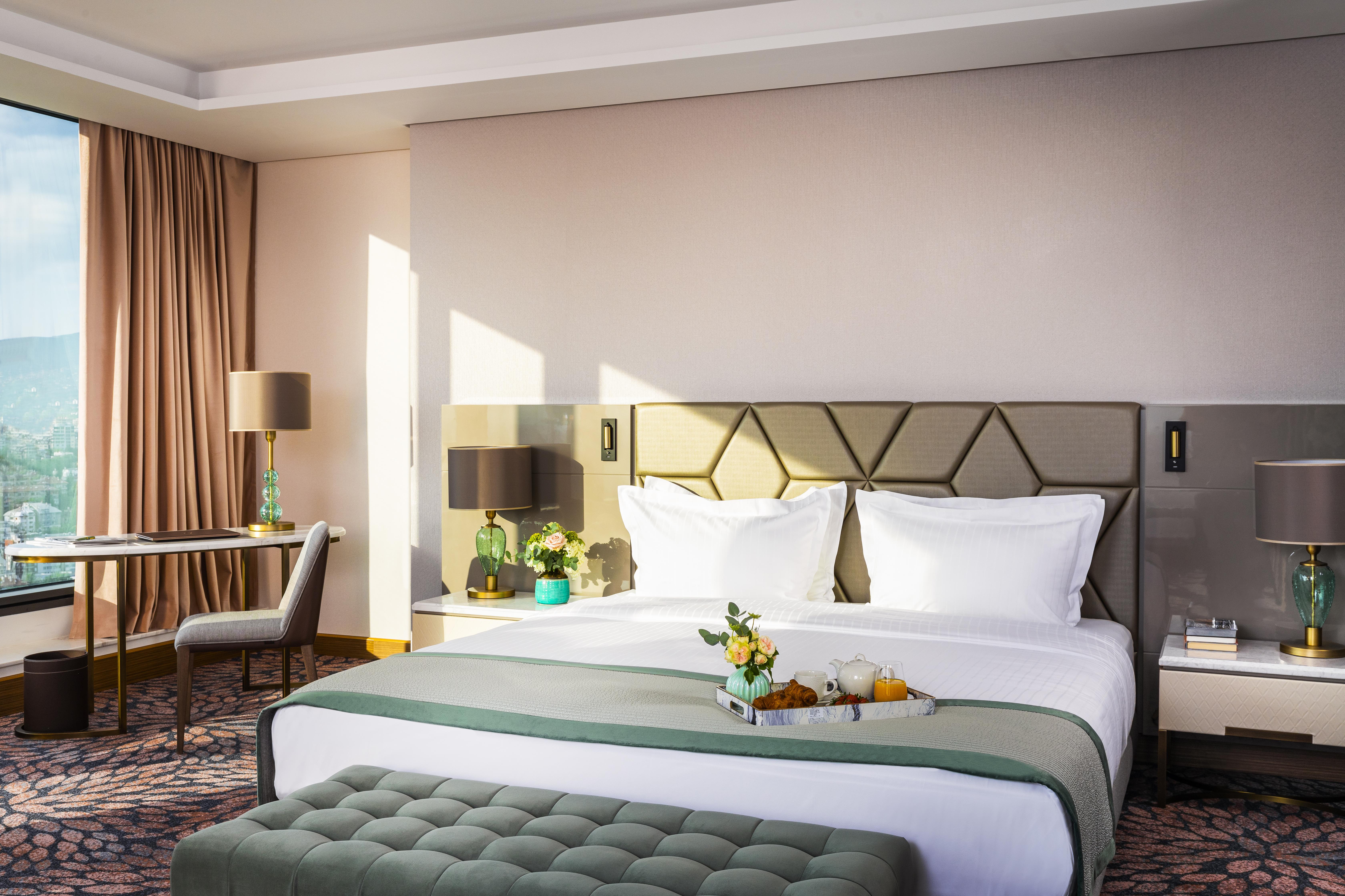 Grand Hotel Millennium โซเฟีย ภายนอก รูปภาพ The photo depicts a stylish hotel room featuring a large bed with white linens and a green decorative throw. Behind the bed is a geometric-patterned headboard. To the side, there are modern bedside lamps and a small table with a vase of flowers. A se