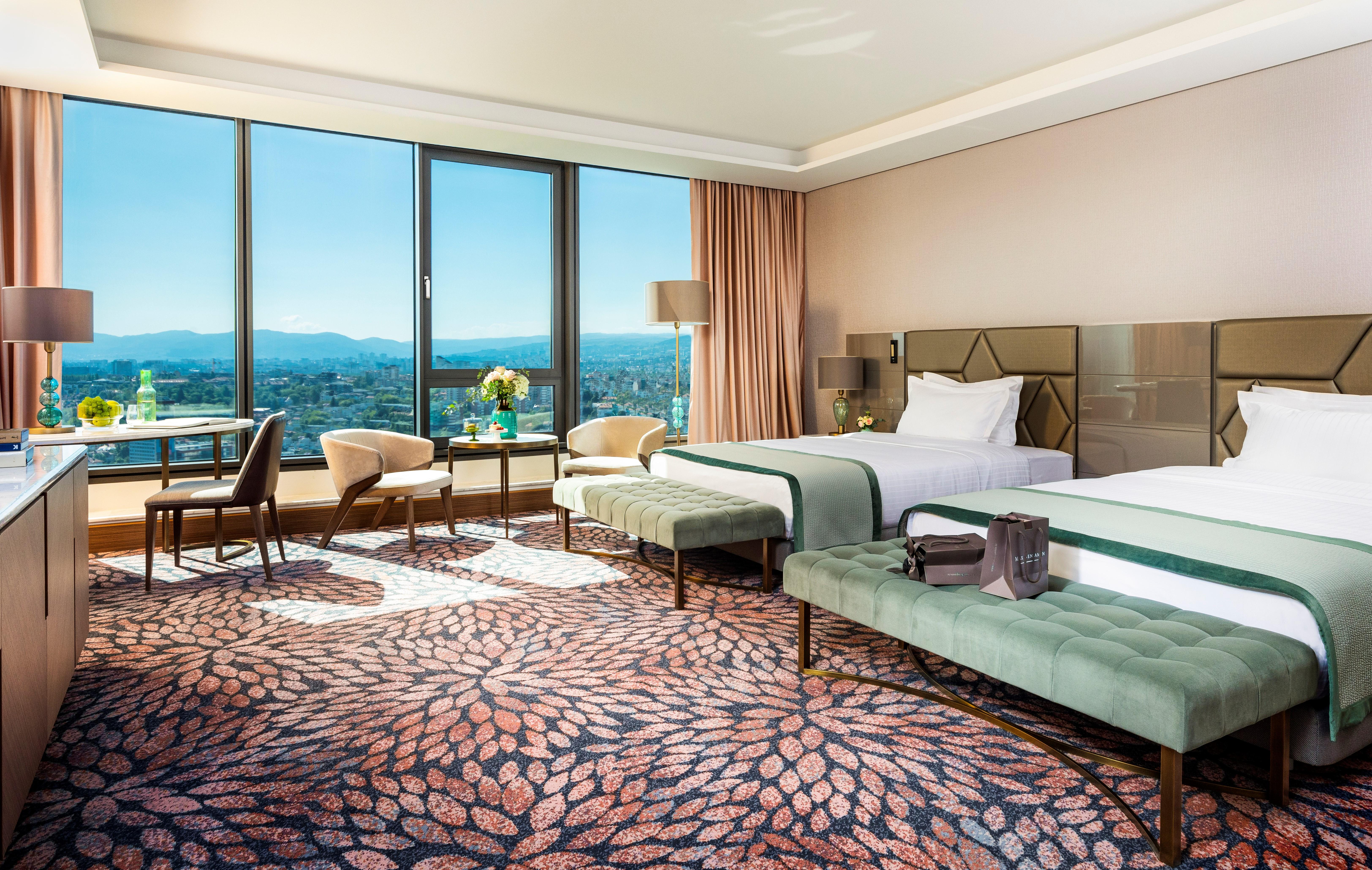 Grand Hotel Millennium โซเฟีย ภายนอก รูปภาพ The photo depicts a luxurious hotel room featuring two beds with elegant bedding. The room has large windows that offer a panoramic view of the city and mountains in the background. There is a decorative area rug with a floral pattern covering the fl