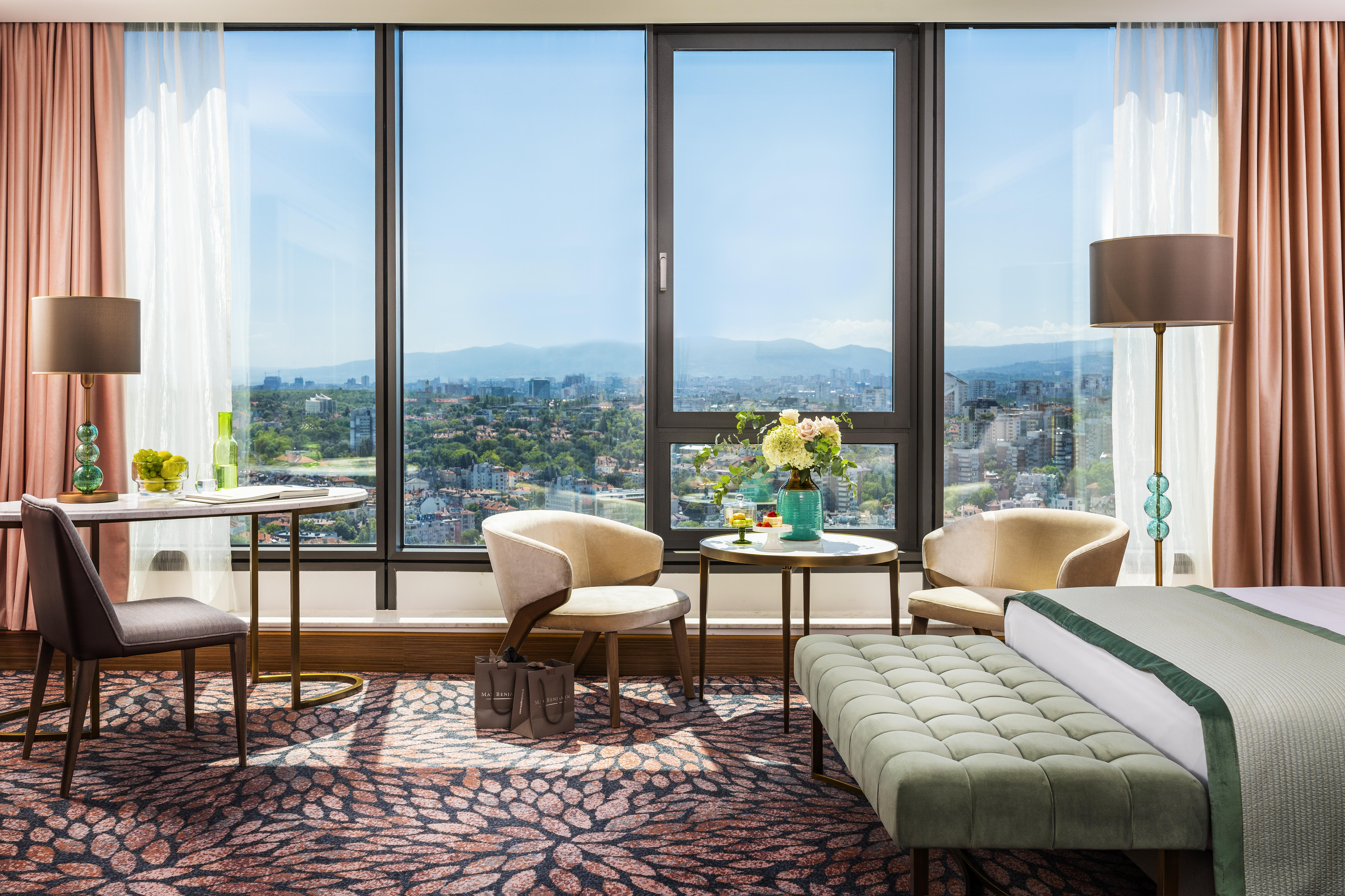 Grand Hotel Millennium โซเฟีย ภายนอก รูปภาพ The photo shows a modern and well-designed hotel room featuring a large window that offers a view of the outdoors, including mountains in the background. Inside the room, there is a neatly made bed with a green accent blanket, a stylish rug with a fl