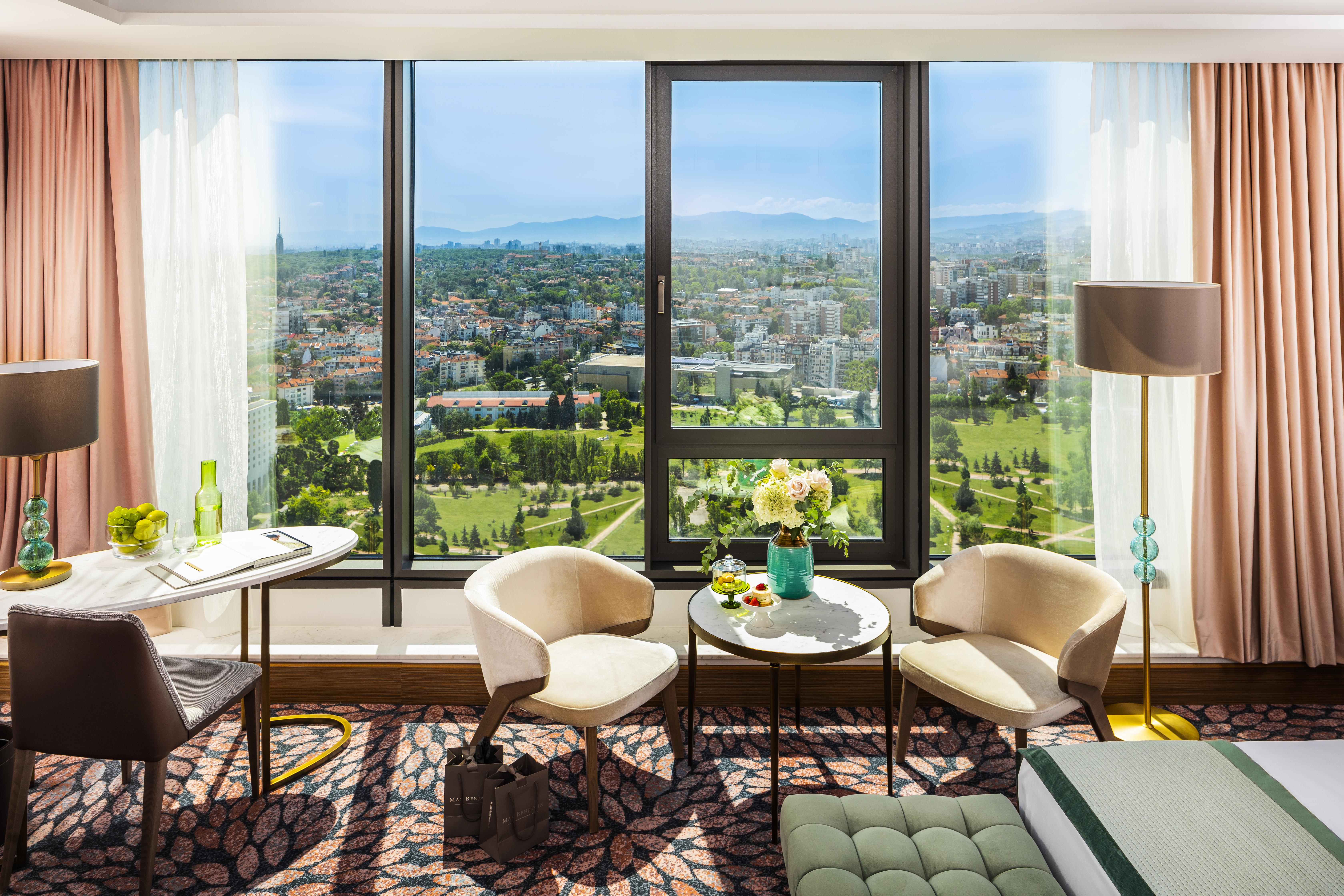 Grand Hotel Millennium โซเฟีย ภายนอก รูปภาพ The photo shows a stylish hotel room with large windows providing a beautiful view of a cityscape and landscape beyond. There are two modern chairs and a small round table set near the windows, accompanied by a vase of fresh flowers. The room feature