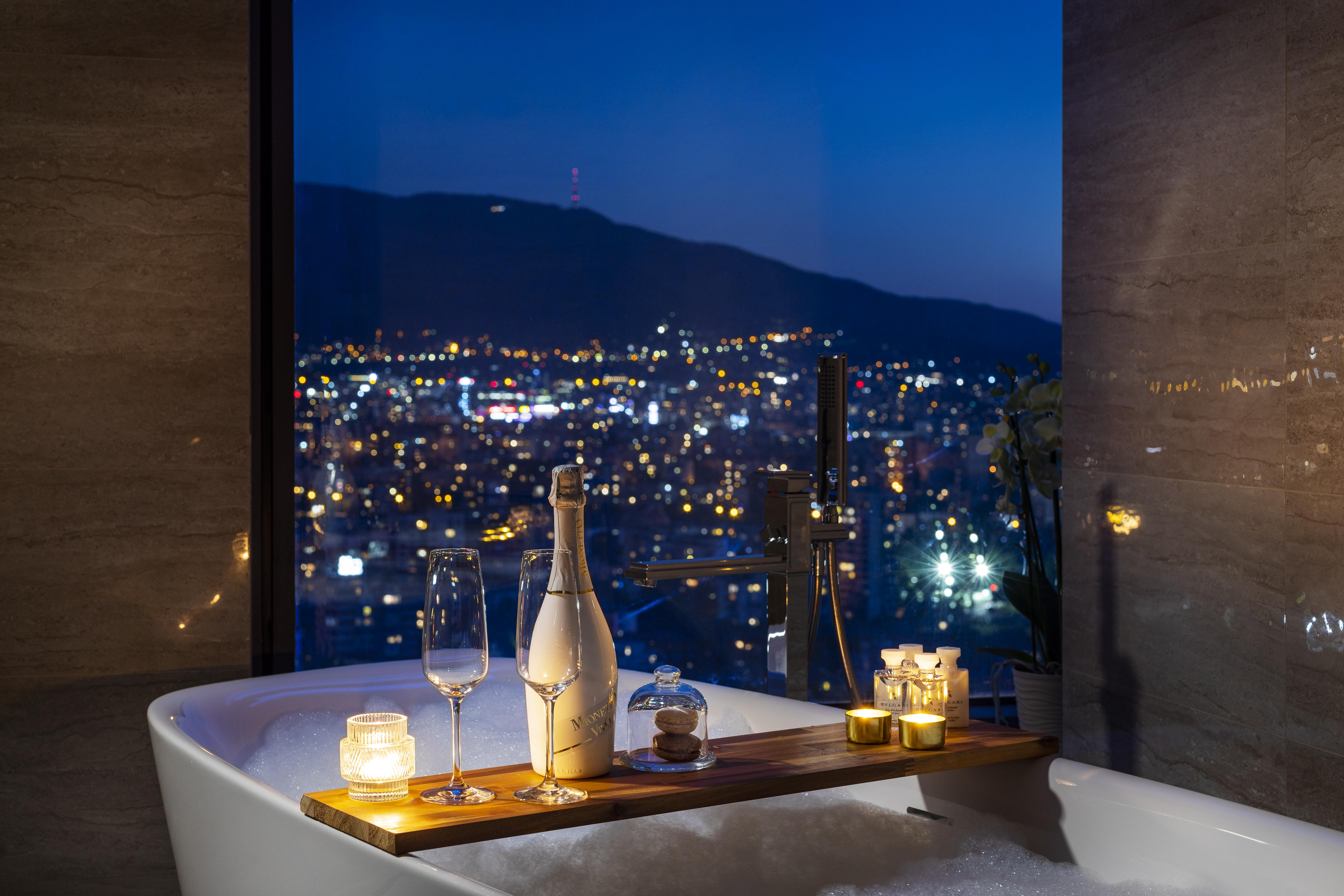 Grand Hotel Millennium โซเฟีย ภายนอก รูปภาพ The photo features a luxurious bathtub placed by a large window, showcasing a scenic city view at night. There is a wooden tray resting on the edge of the tub, displaying a bottle of champagne, two glass flutes, and several lit candles, creating an a