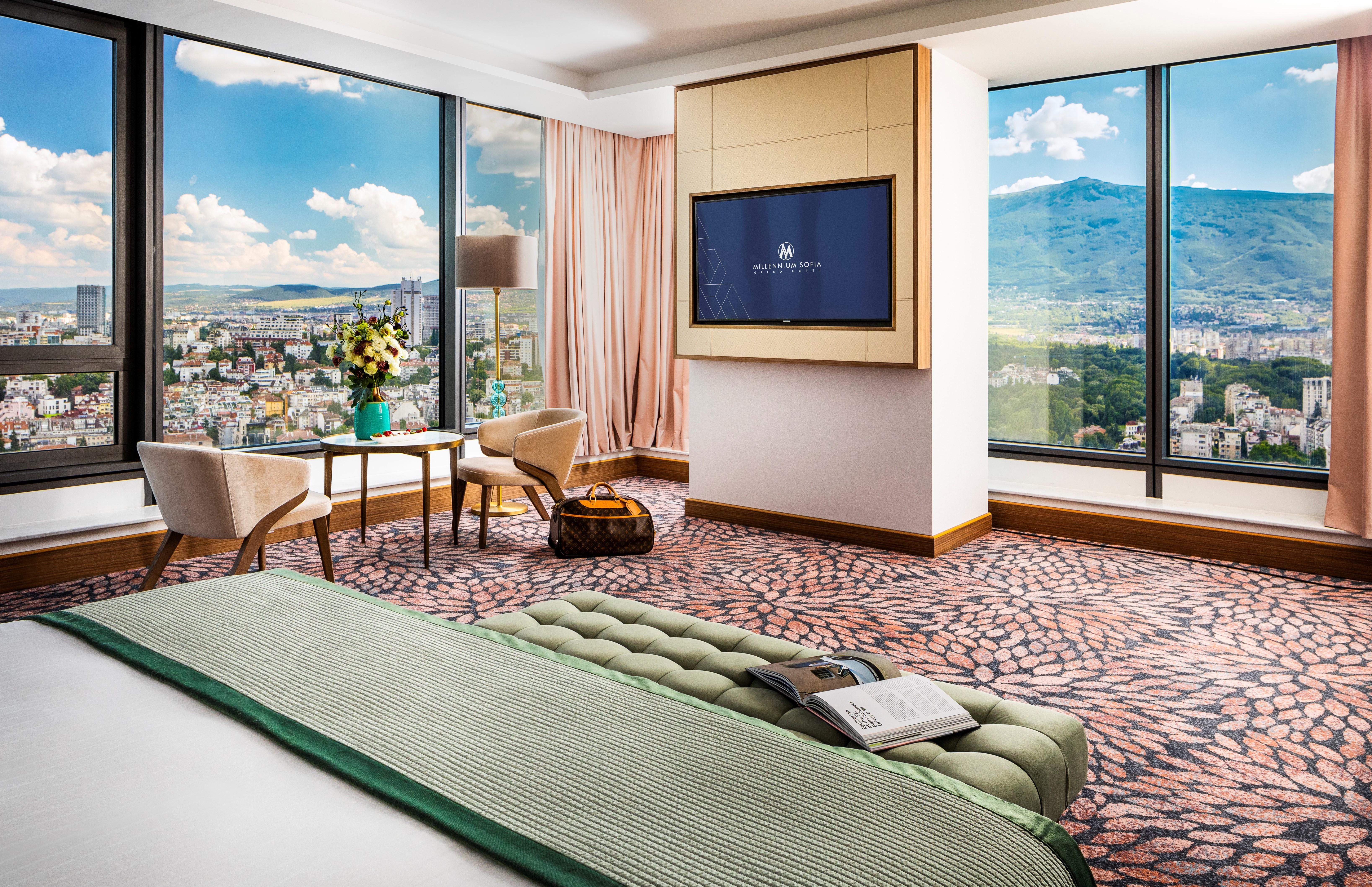 Grand Hotel Millennium โซเฟีย ภายนอก รูปภาพ The photo showcases a stylish hotel room with modern decor. It features large windows providing a panoramic view of a cityscape and mountains in the background. Inside, there is a comfortable bed with a green bedspread, a seating area with two armcha
