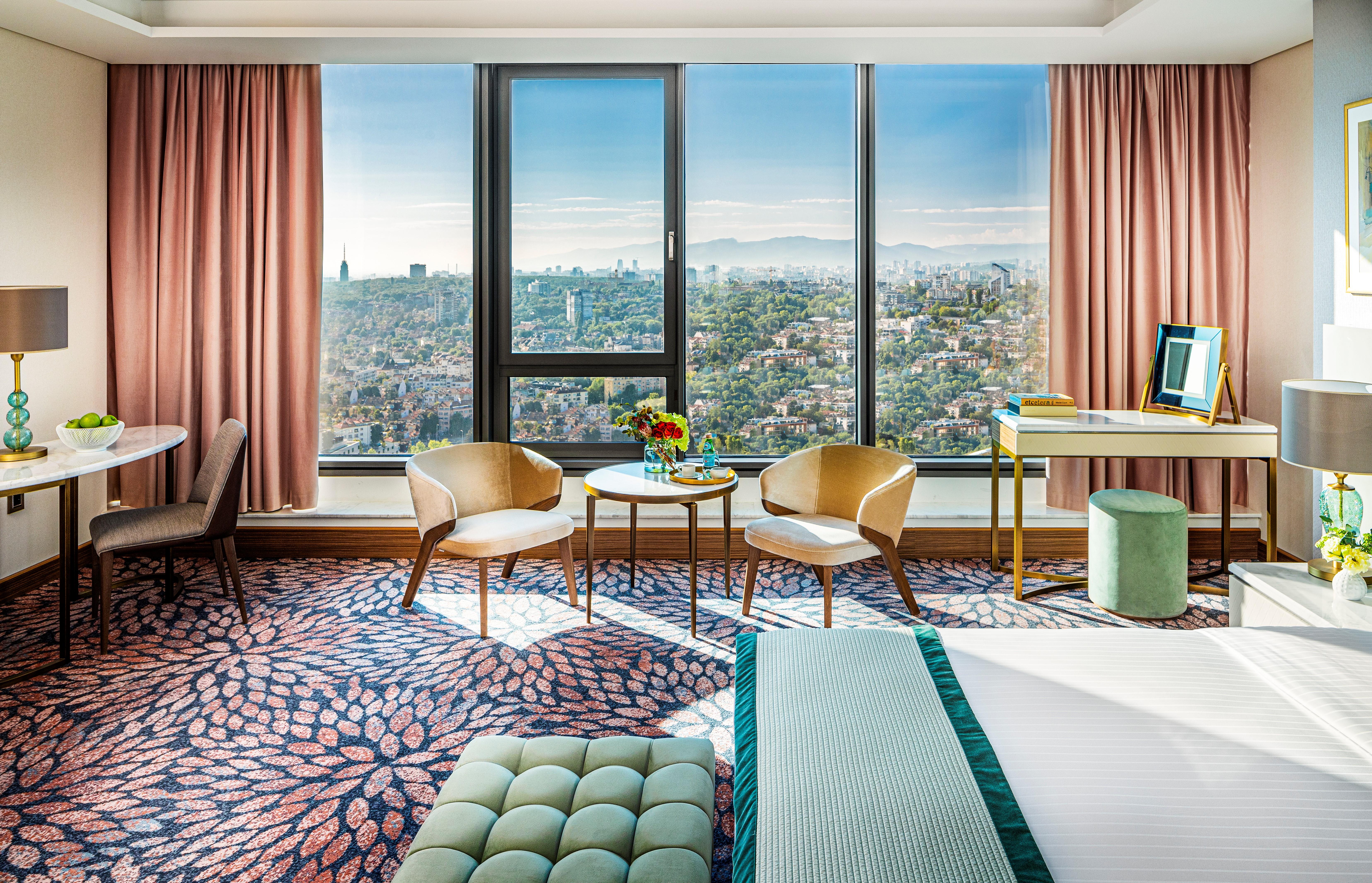 Grand Hotel Millennium โซเฟีย ภายนอก รูปภาพ The photo shows a stylish hotel room featuring modern decor. There are large windows that offer a panoramic view of the cityscape, allowing natural light to fill the space. The walls are adorned with soft pink curtains. In the room, there are two ele