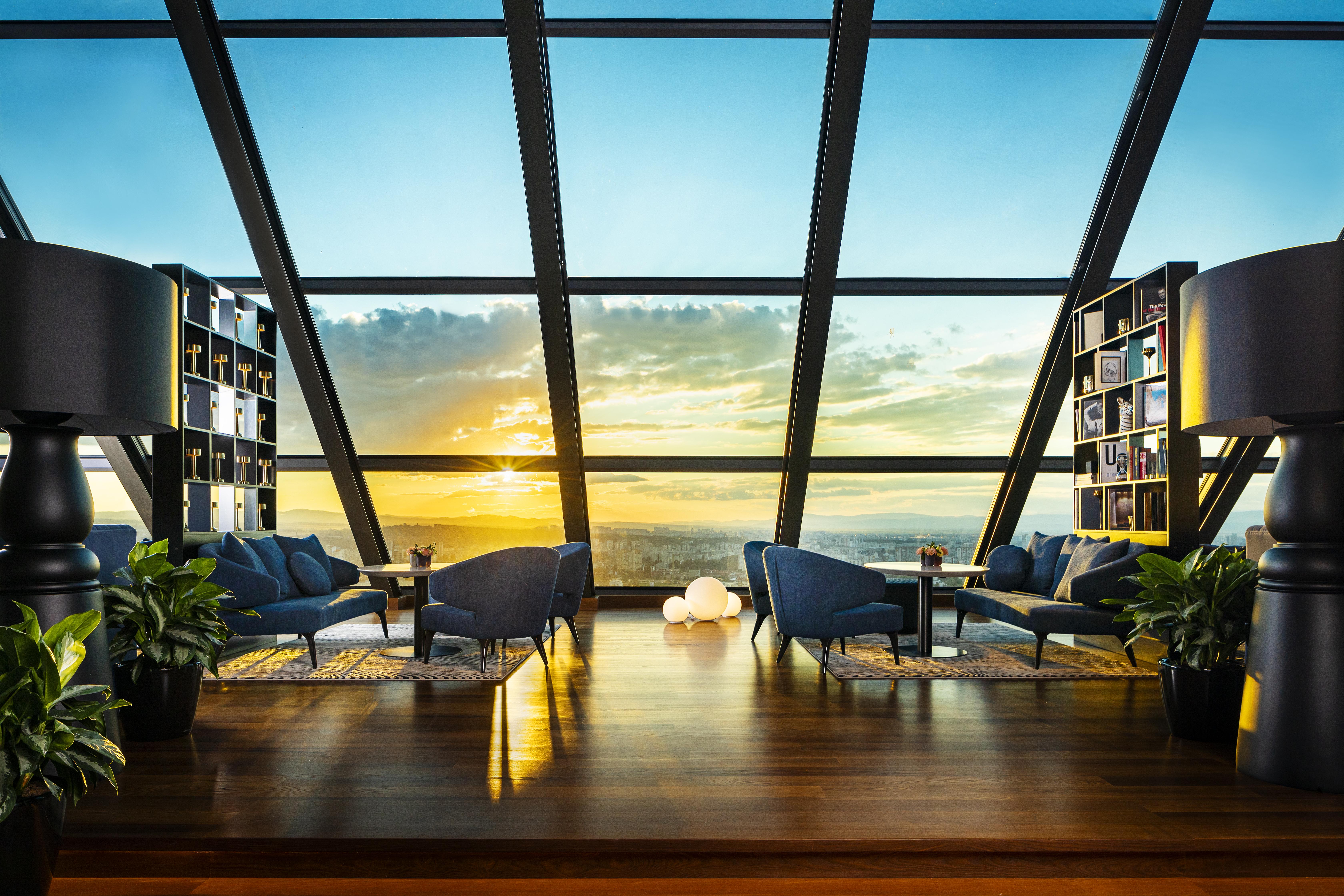 Grand Hotel Millennium โซเฟีย ภายนอก รูปภาพ The photo depicts a modern, stylish interior of a high-rise space with a panoramic view through large glass windows. The scene features a cozy seating area with several blue armchairs arranged around a low table, creating a comfortable atmosphere. Th