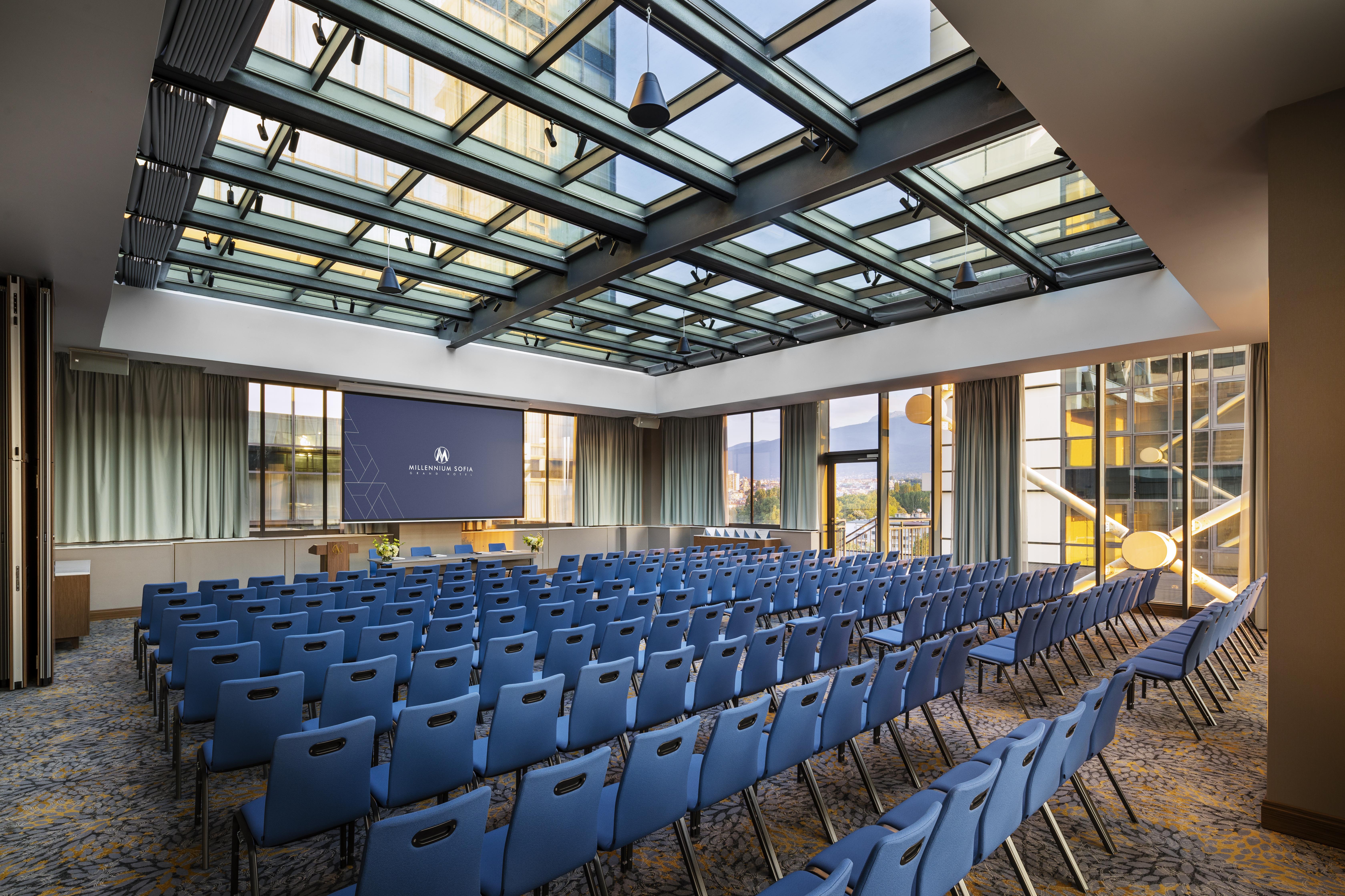 Grand Hotel Millennium โซเฟีย ภายนอก รูปภาพ The photo shows a large conference room or meeting space. It features rows of blue chairs arranged in a theater style, facing a large screen at the front of the room. The room is well-lit, with a glass ceiling allowing natural light to filter in. The