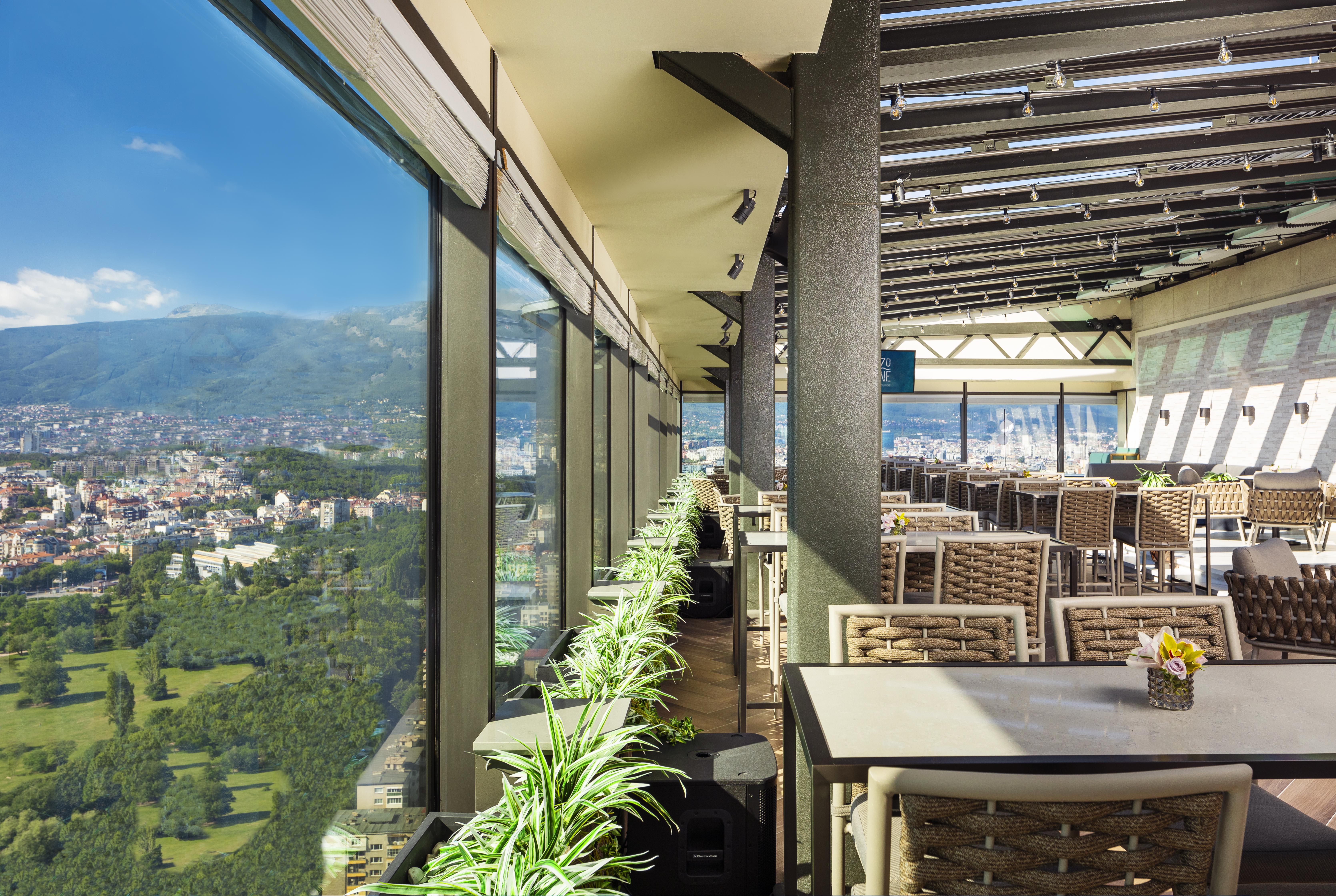 Grand Hotel Millennium โซเฟีย ภายนอก รูปภาพ The photo shows a modern, spacious restaurant with large glass windows offering a panoramic view of a city and surrounding landscape. The interior features contemporary furniture, including stylish tables and chairs. There are potted plants along the